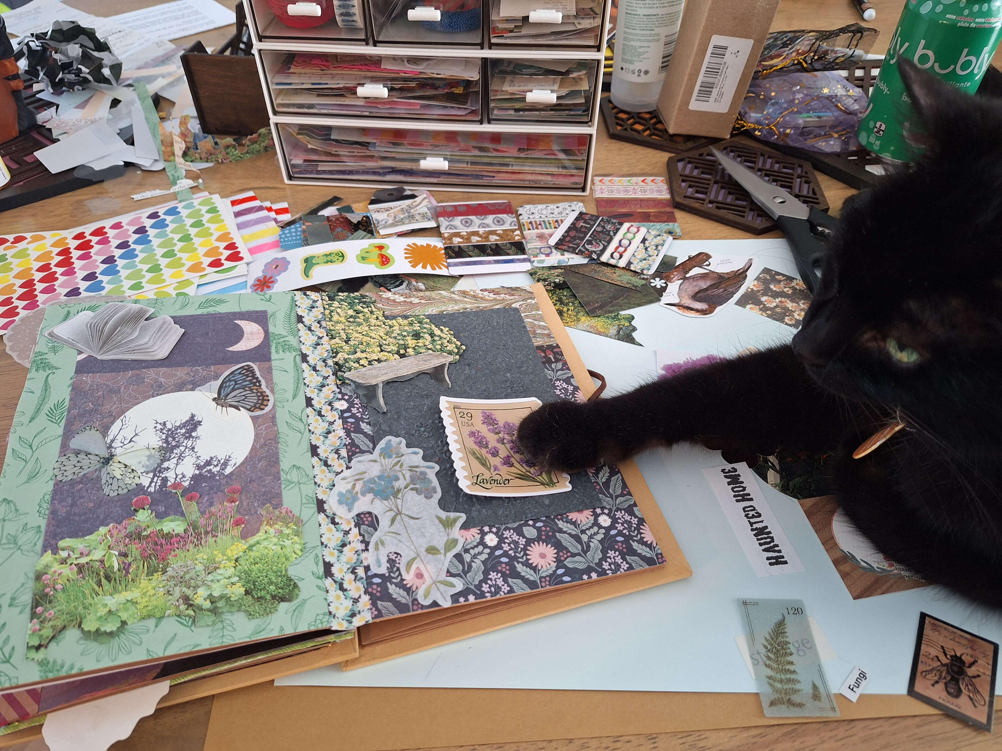 Chesty helps A black cat lies on a table covered in craft supplies, her paw placed delicately on top of a sticker that has yet to be added to a scrapbook.